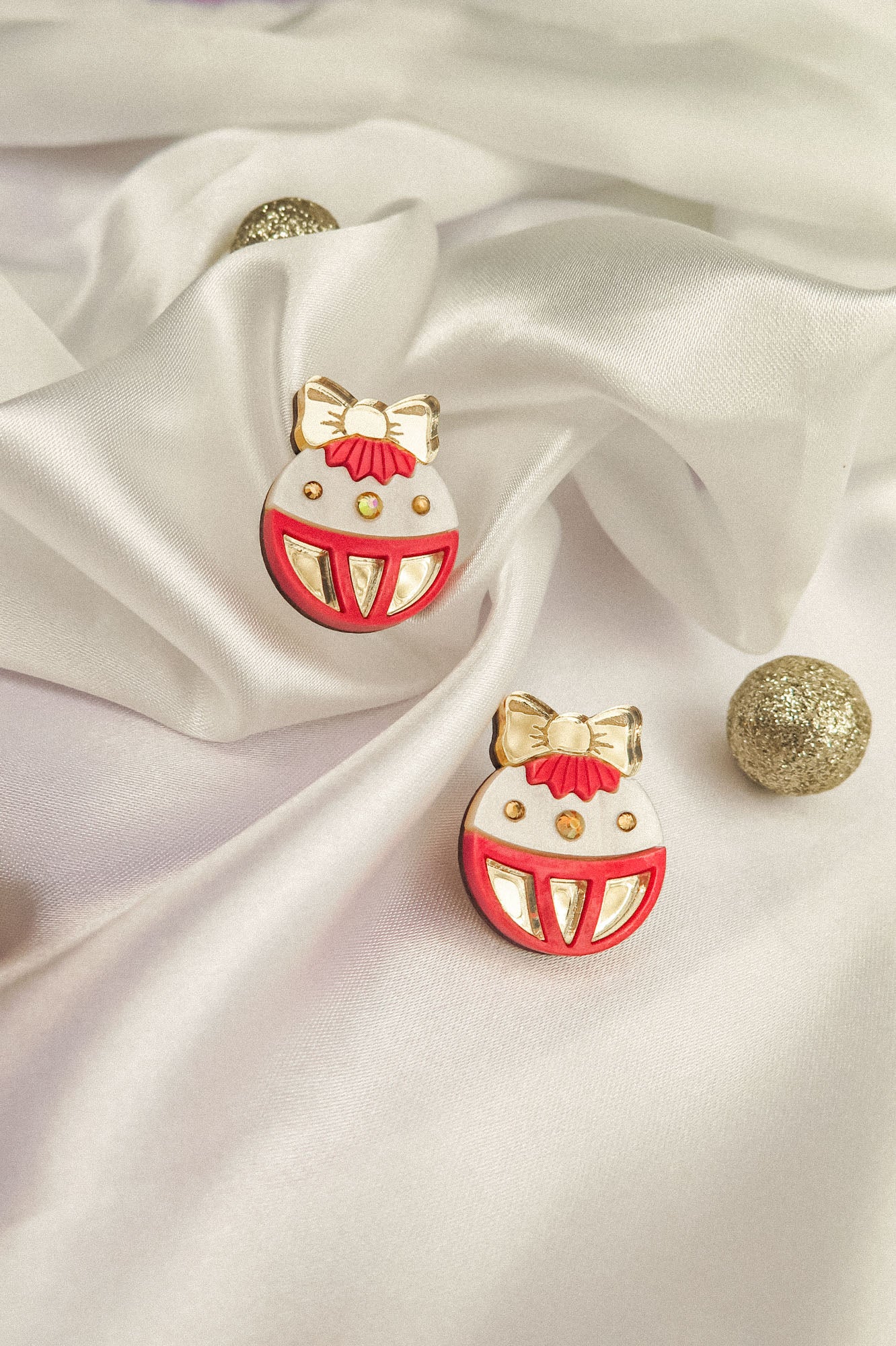 A pair of round enamel pins with red, white, and gold details, resembling festive ornaments with bows, are displayed on white satin fabric with gold glittery balls—perfect as holiday jewelry or a festive earrings-inspired accent.