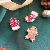 Three decorated cookies shaped like a mitten, a stocking, and a gingerbread person rest on green fabric with gold ribbon, glittery ornaments, pinecones, and a handcrafted accessory to complete the festive holiday style.