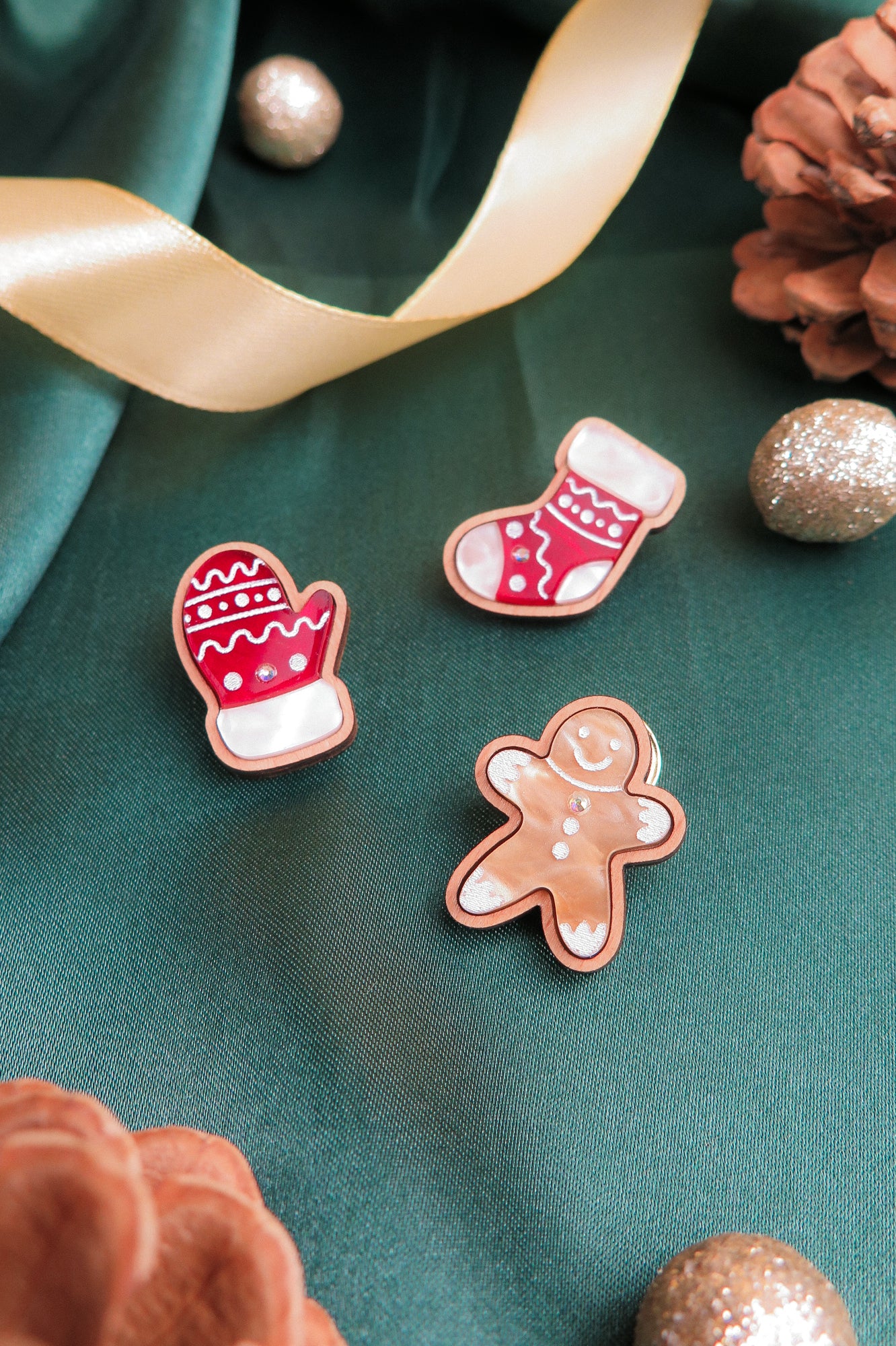 Three handcrafted Christmas cookies—a mitten, a stocking, and a gingerbread person—sit on green fabric with a gold ribbon, pinecones, and sparkly ornaments, adding festive charm to your holiday wardrobe.