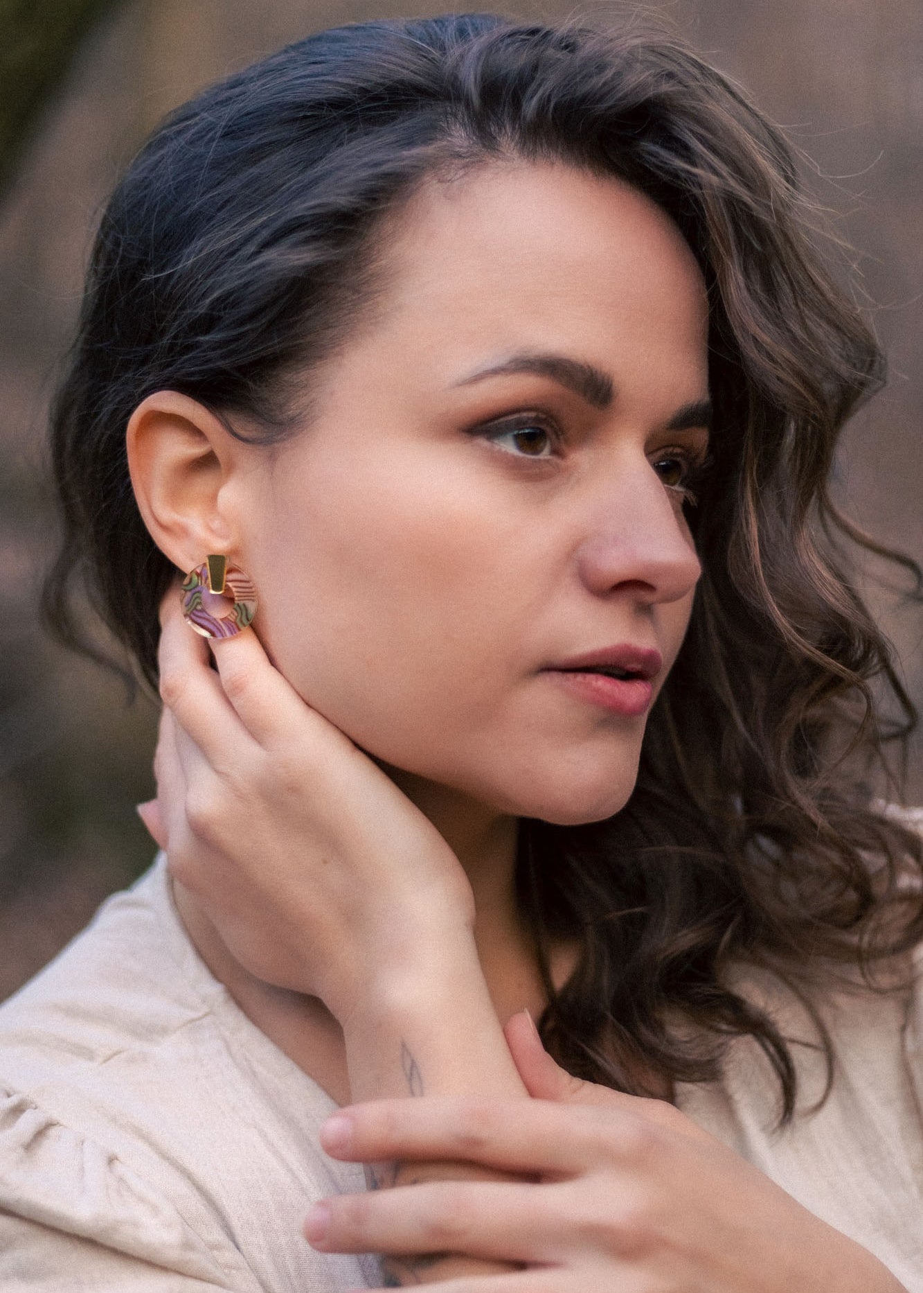 A woman with wavy brown hair wearing a beige top touches her Minimalist Circle Stud Earrings with one hand and rests her other hand on her collarbone. She looks slightly to the side with a calm expression. The earrings are hypoallergenic.