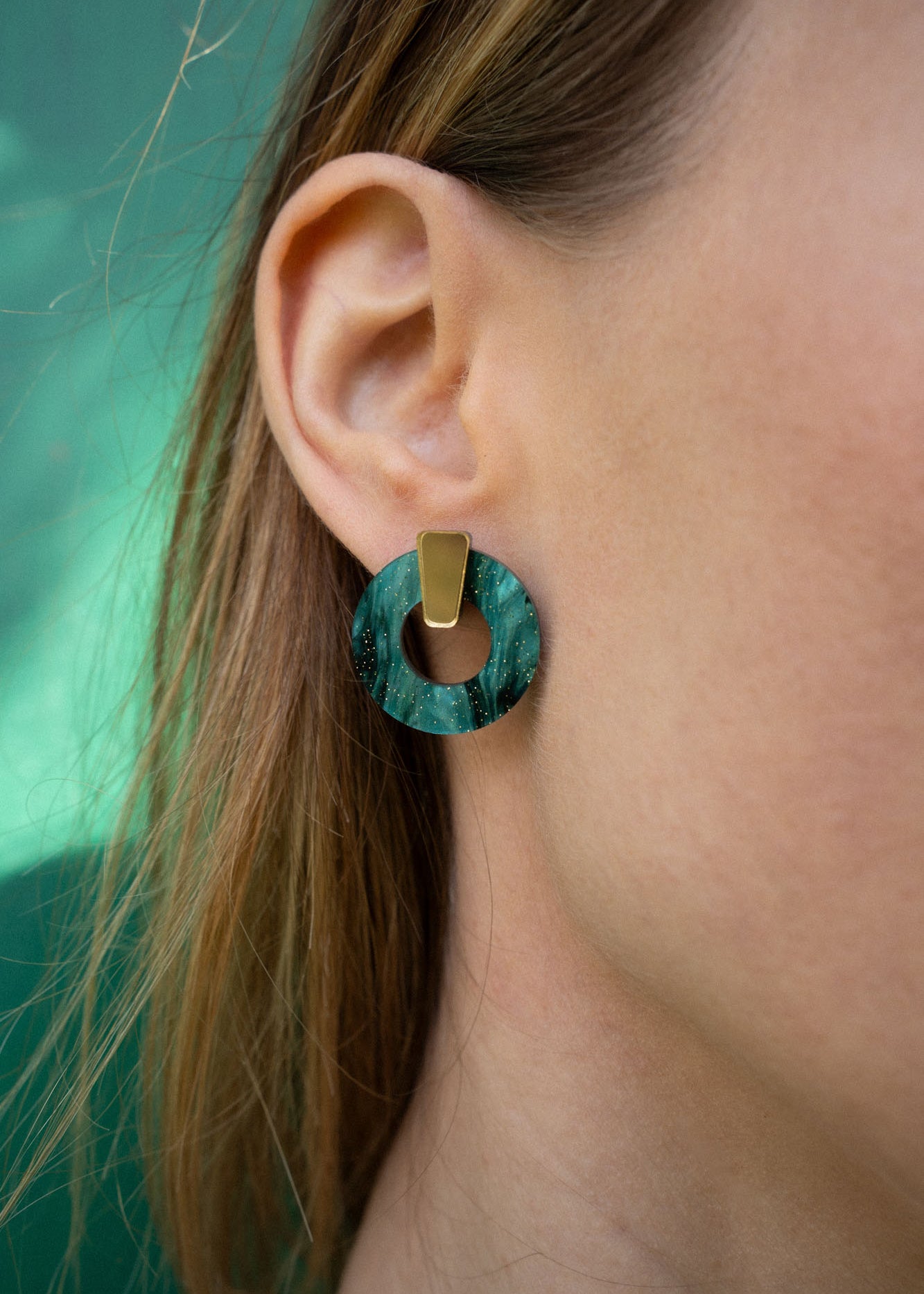 Close-up of a womans ear wearing a hypoallergenic, dark green marbled Minimalist Circle Stud Earring with a gold rectangular accent; her light brown hair partially frames her ear.