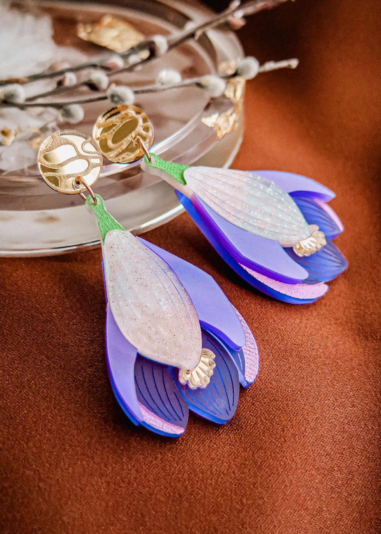 A pair of handcrafted Crocus Flower Earrings, shaped like purple and white blossoms with gold circular studs, rest on a brown surface near a glass dish and decorative twig—a stunning statement accessory.