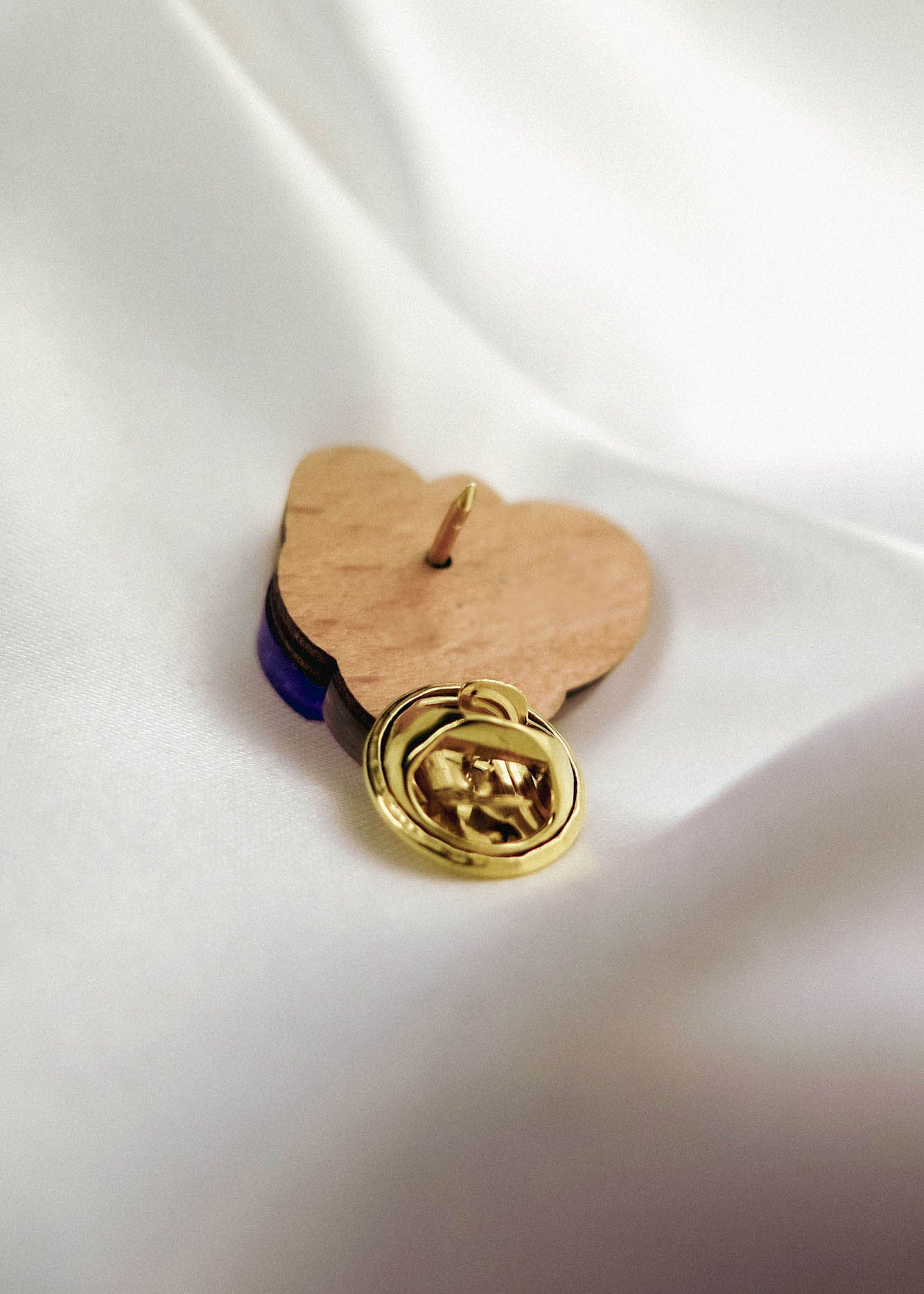 A handcrafted gold lapel pin with an embossed hand design rests on a wooden heart-shaped base, placed on a smooth white fabric background—an ideal gift for nature lovers.
