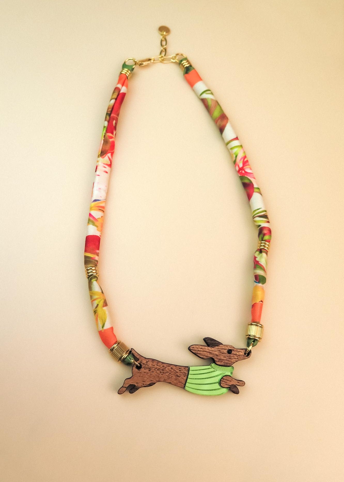 A colorful handcrafted Dachshund necklace featuring a patterned fabric chain and a pendant of a running dachshund in a green sweater. Made with recycled materials, it boasts gold details against a light beige background.