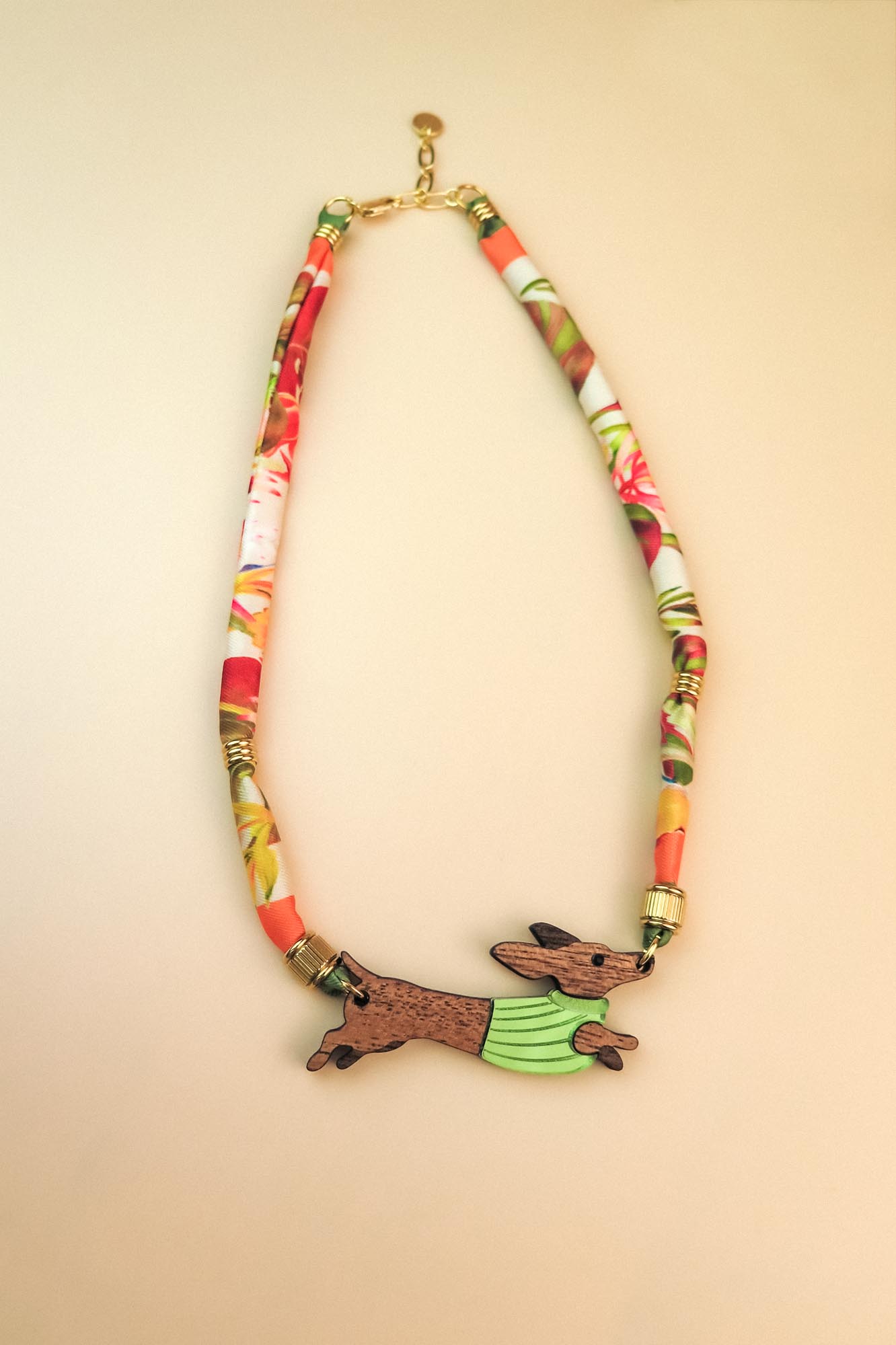 A colorful handcrafted Dachshund necklace featuring a patterned fabric chain and a pendant of a running dachshund in a green sweater. Made with recycled materials, it boasts gold details against a light beige background.