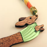 A close-up of a handcrafted wooden keychain shaped like a dog in a green sweater, attached to a colorful, twisted cord with a gold-colored metal clasp.