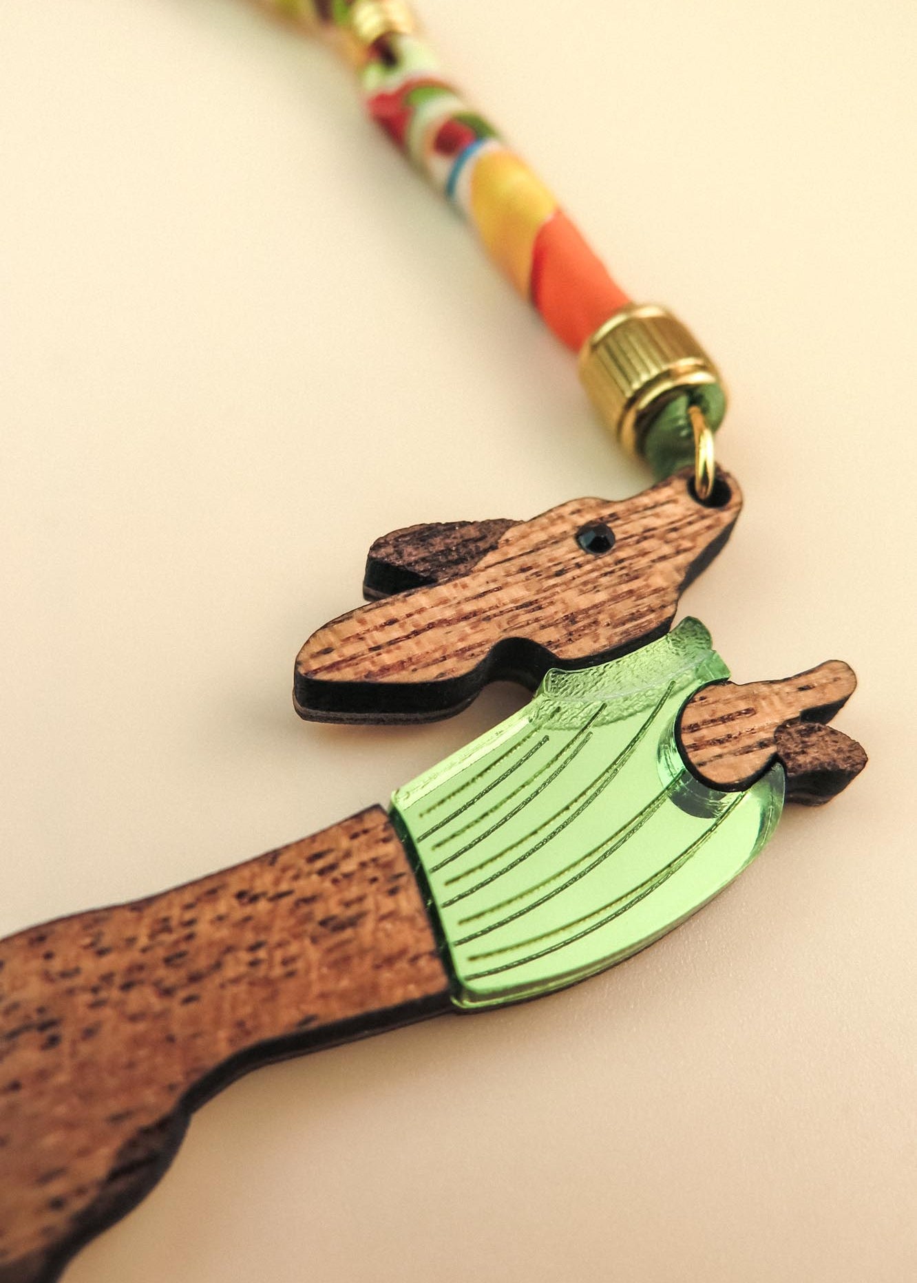 A close-up of a handcrafted wooden keychain shaped like a dog in a green sweater, attached to a colorful, twisted cord with a gold-colored metal clasp.