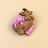 A wooden Dachshund brooch shaped like a brown dog lying on its back on a pink pillow with small decorative marks, set against a plain beige background—perfect for playful jewelry lovers.