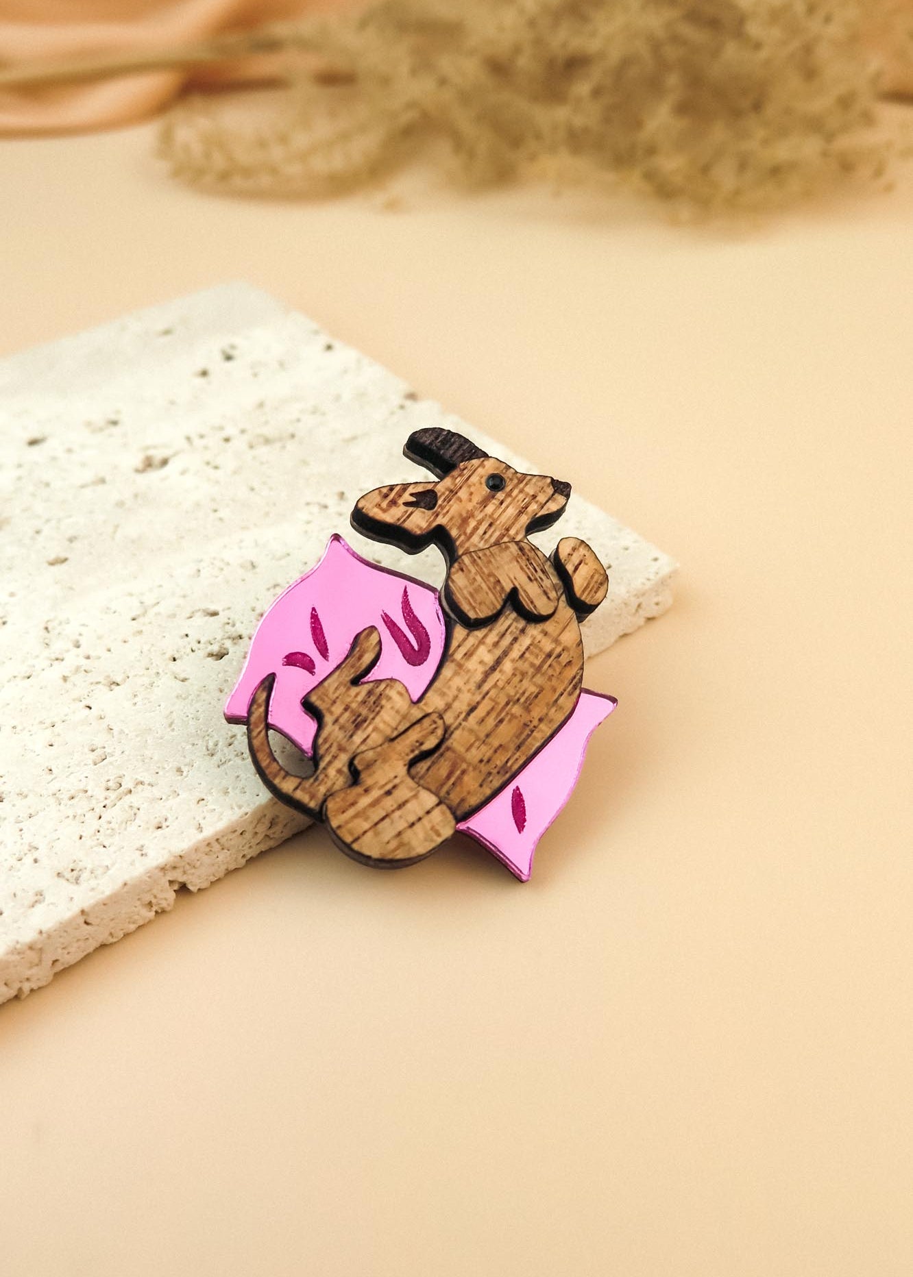 A playful Dachshund brooch shaped like a curled-up dog holding a pink, leaf-shaped object rests on a textured white stone slab against a soft beige background.