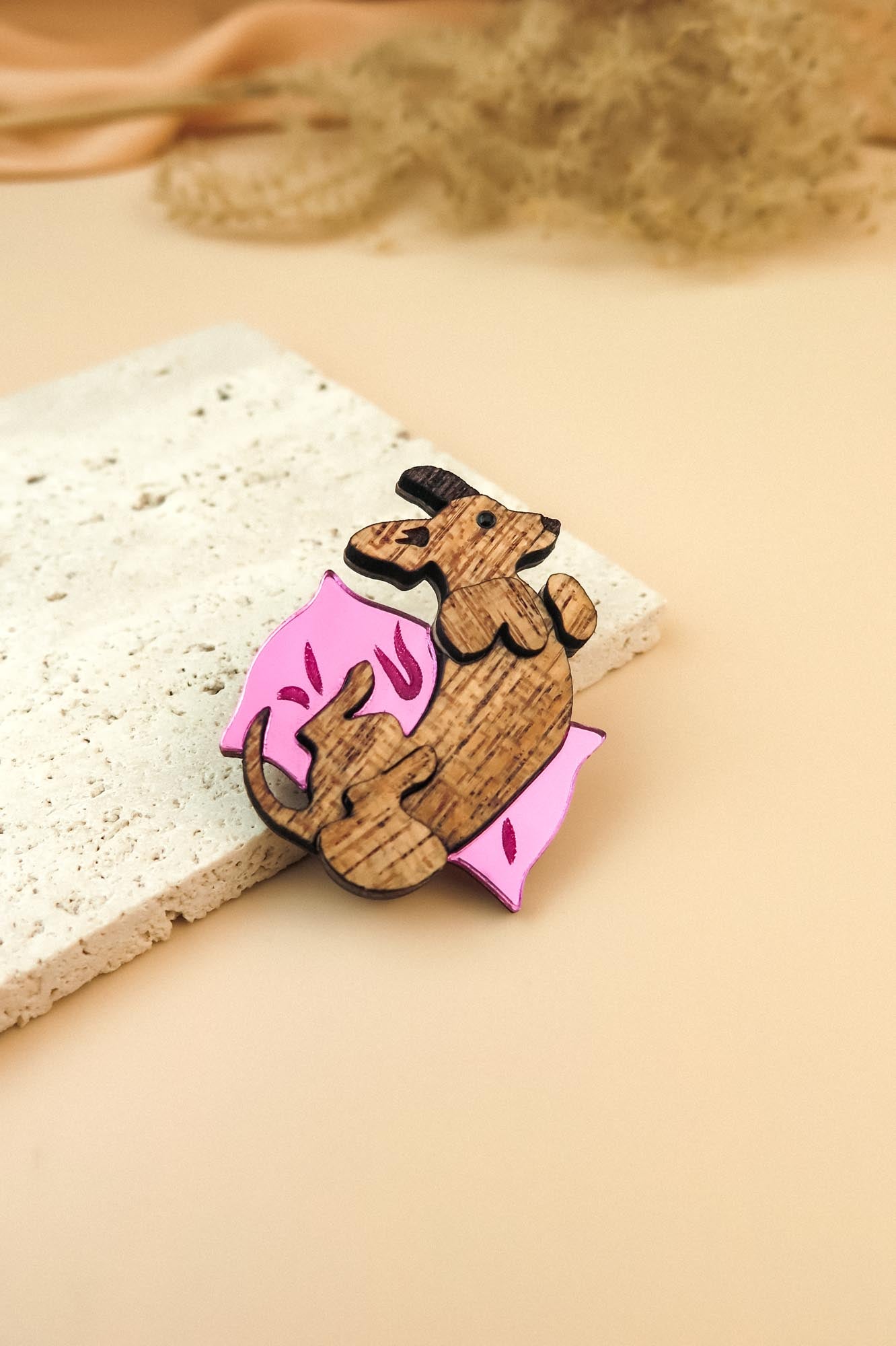 A playful Dachshund brooch shaped like a curled-up dog holding a pink, leaf-shaped object rests on a textured white stone slab against a soft beige background.