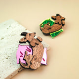 Two wooden pins shaped like sleeping cats, one lying on a pink pillow with leaf designs and the other on a green pillow. Both are displayed as playful jewelry on a light beige surface next to a textured stone.