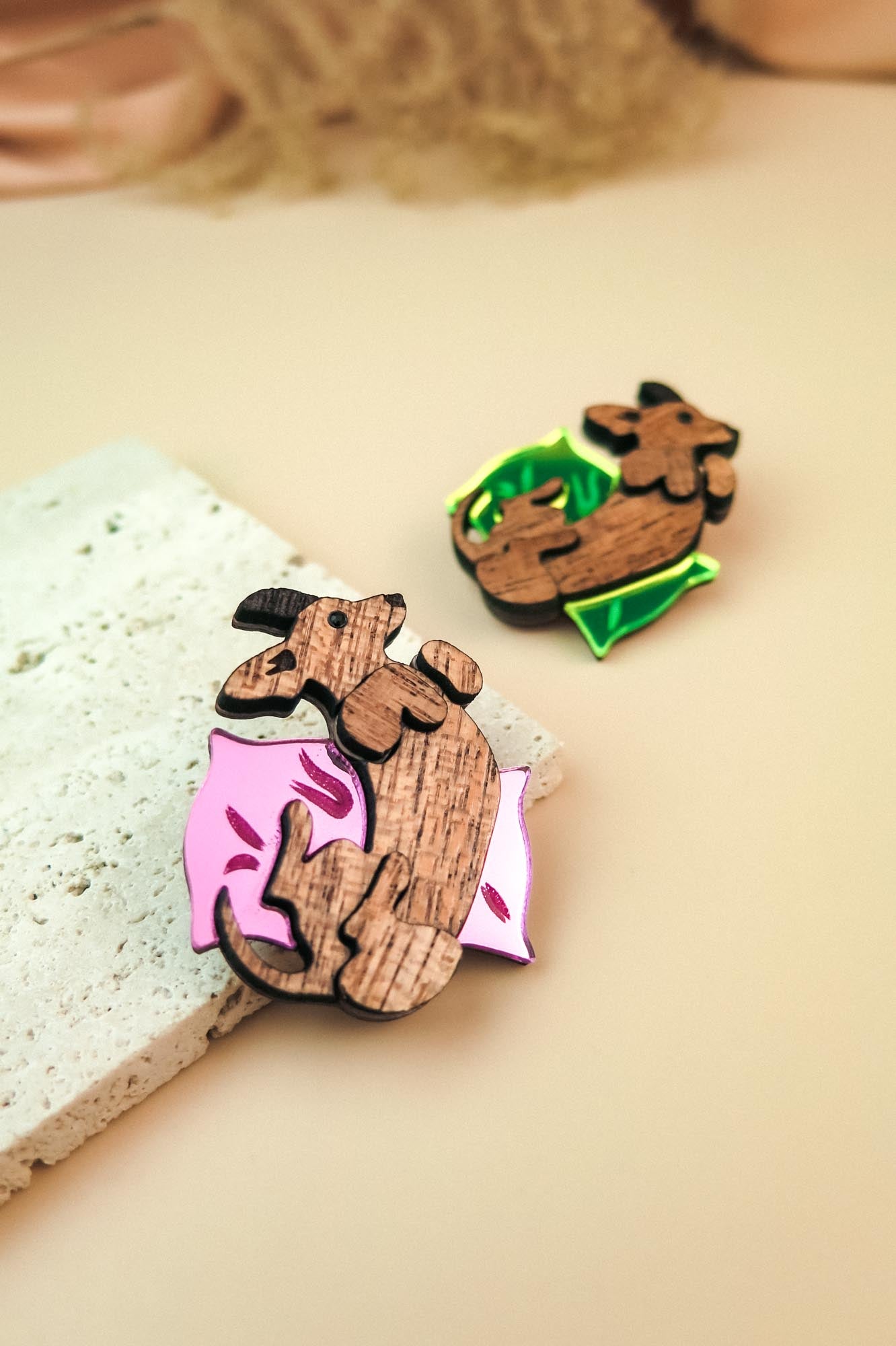 Two wooden pins shaped like sleeping cats, one lying on a pink pillow with leaf designs and the other on a green pillow. Both are displayed as playful jewelry on a light beige surface next to a textured stone.