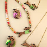 Colorful jewelry set featuring dachshund-shaped necklaces and brooches; the dogs wear green or pink colorful sweaters, with beads and gold chain accents, crafted from sustainable wood and arranged on a neutral beige background.