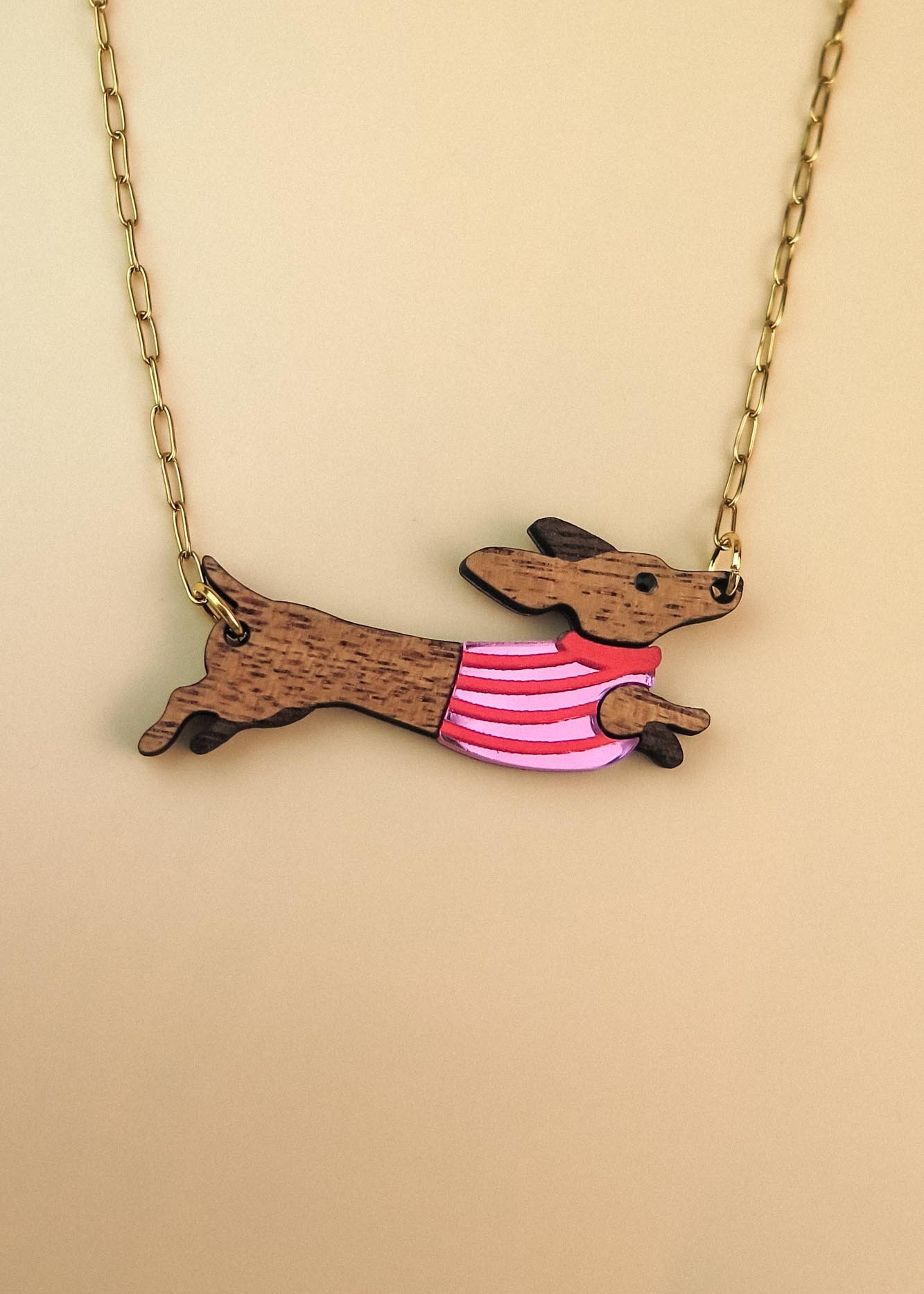 Gold chain necklace with a pendant shaped like a brown Dachshund dog, wearing a colorful sweater and crafted from sustainable wood, set against a beige background.