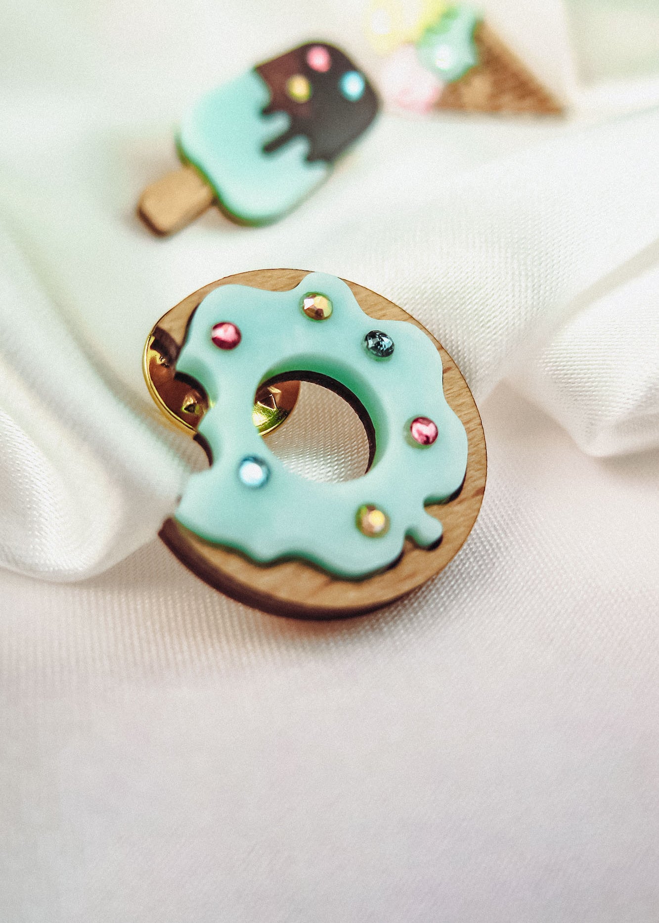 A handcrafted wooden Donut Pin with light blue icing and sparkling rhinestones rests on white fabric. In the background, blurred pins shaped like an ice cream cone and a popsicle add a playful touch.