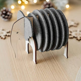 A wooden and felt elephant coasters holder sits on a light table, adding witty design and furniture protection, with pinecones, snowflake decorations, and string lights blurred in the background.