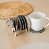 A set of round Elephant Coasters with witty design sits in a wooden elephant-shaped holder on a table, offering furniture protection while a white mug rests on one coaster.