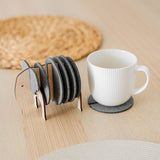 A white ribbed coffee cup sits on a round gray coaster. Beside it, Elephant Coasters with witty design are stacked in a wooden elephant-shaped holder, providing both style and furniture protection on the light wooden table. A woven placemat is in the background.