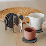 A set of Elephant Coasters with witty design offers stylish furniture protection, nestled in a wooden elephant-shaped holder on a wooden table beside a pink ceramic cup, white mug, and woven placemat.
