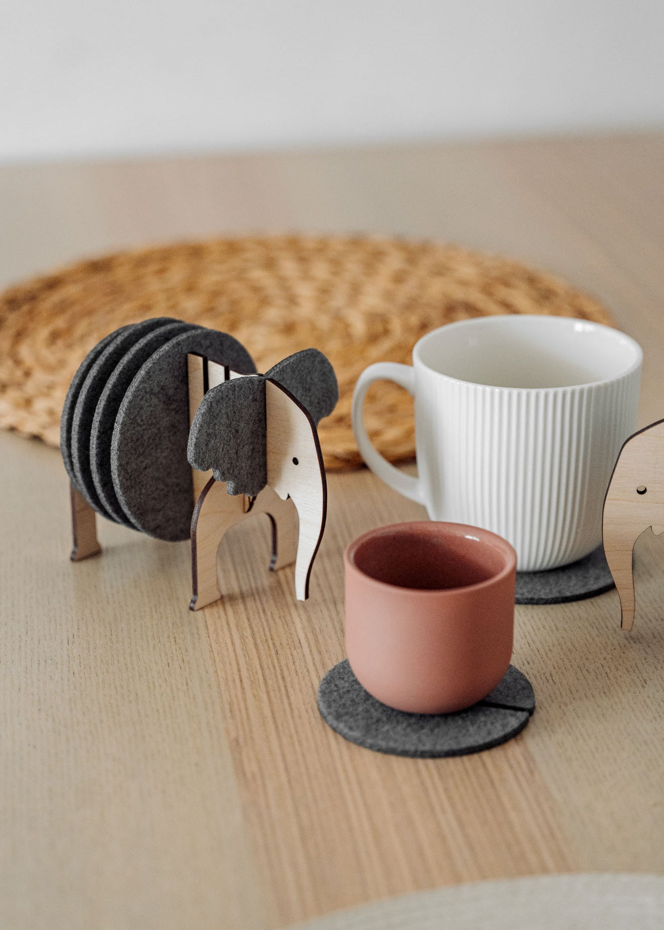 A set of Elephant Coasters with witty design offers stylish furniture protection, nestled in a wooden elephant-shaped holder on a wooden table beside a pink ceramic cup, white mug, and woven placemat.