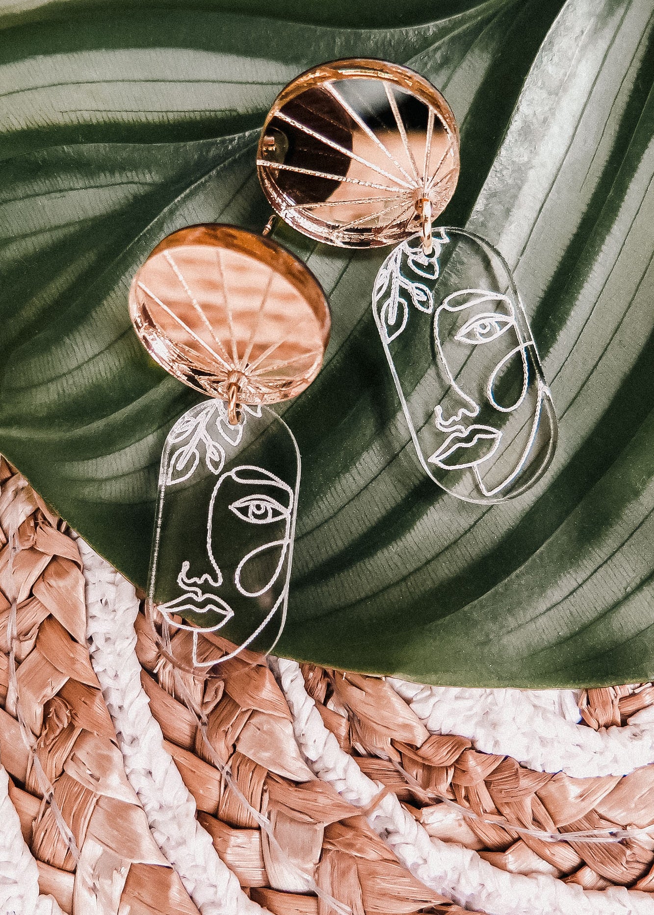Gold and clear handcrafted Face Earrings with abstract faces rest on a large green leaf atop a woven straw mat, adding artful sophistication to elevate your style.