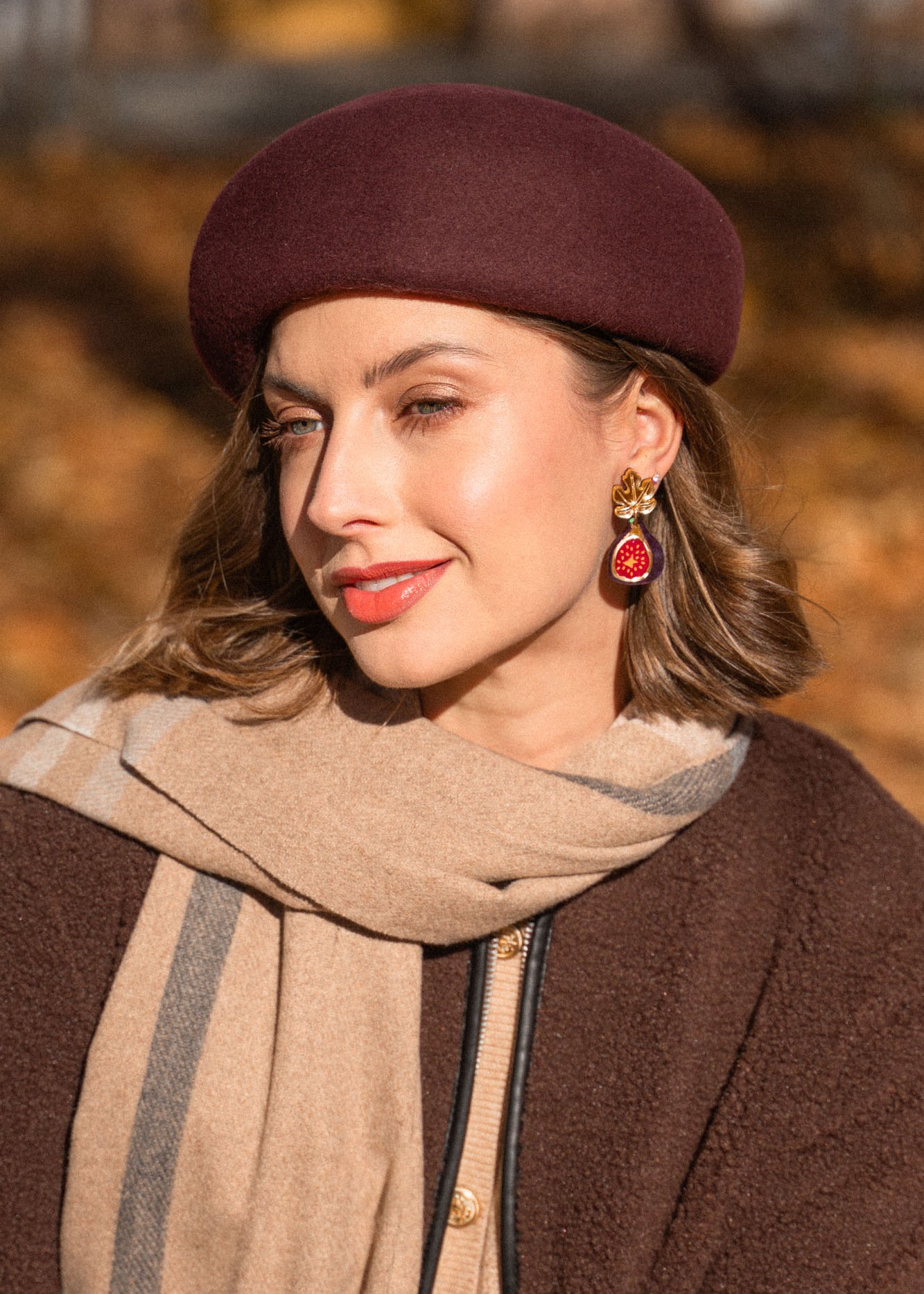 A woman wearing a maroon beret, beige and brown scarf, and matching coat smiles softly outdoors. She has light makeup and wears handcrafted Fig Earrings with gold mirror leaves. The background is blurred with autumn colors.