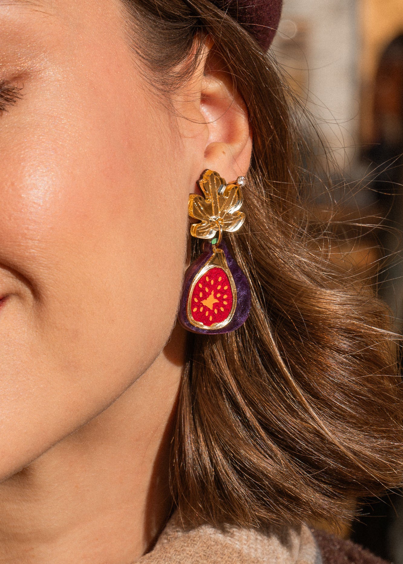 A woman outdoors smiles gently with her eyes closed. She wears a burgundy beret, a tan scarf with a fig brooch, and handcrafted fig earrings accented with gold mirror leaves. Autumn trees and buildings are blurred in the background.