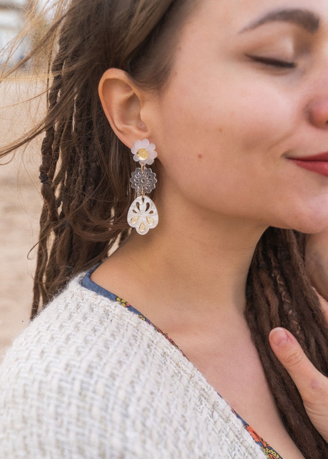 A woman with closed eyes and long brown dreadlocks wears a beige sweater and large handcrafted boho earrings, standing outdoors on a sandy beach. She smiles softly and touches her chest with one hand.