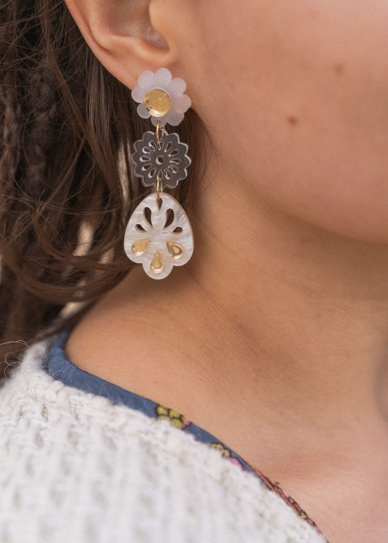 A close-up of a person wearing handcrafted, dangling earrings with flower and abstract shapes in translucent and gold tones, paired with a white textured sweater and a blue shirt with a floral pattern.