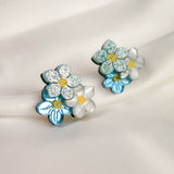 A pair of Forget Me Not Studs, featuring blue and white petals, yellow centers, and glittery accents, these floral earrings are true wearable art displayed on a soft, light-colored fabric background.