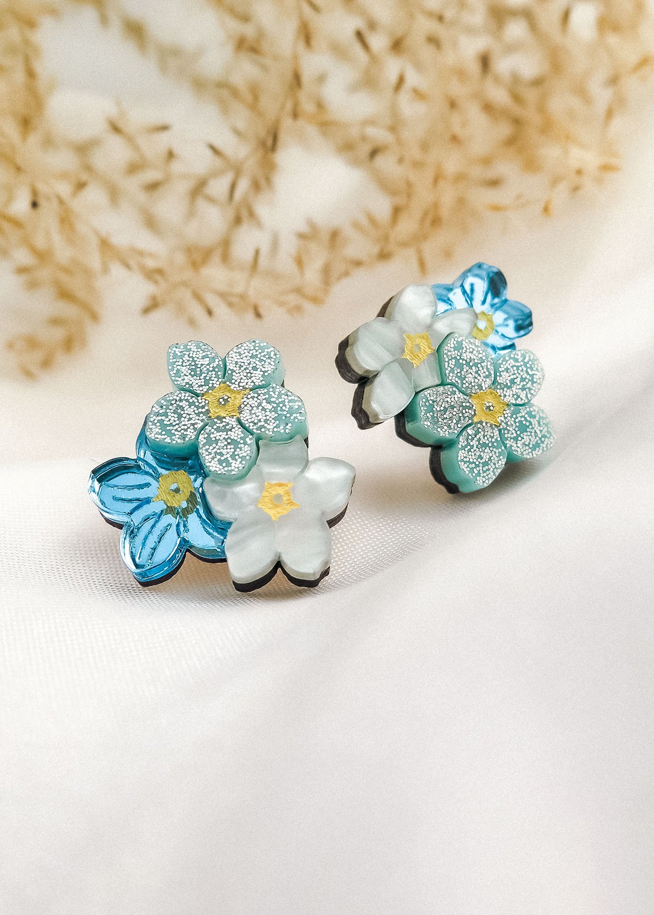 A pair of decorative Forget Me Not Studs featuring layered blue and white flowers with glittery petals, displayed on a soft white fabric with dried plants in the background—delicate floral earrings that are true wearable art.