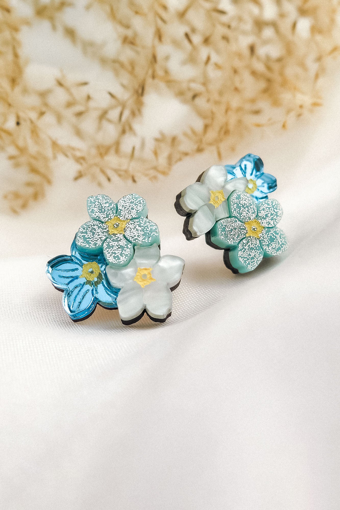 A pair of decorative Forget Me Not Studs featuring layered blue and white flowers with glittery petals, displayed on a soft white fabric with dried plants in the background—delicate floral earrings that are true wearable art.