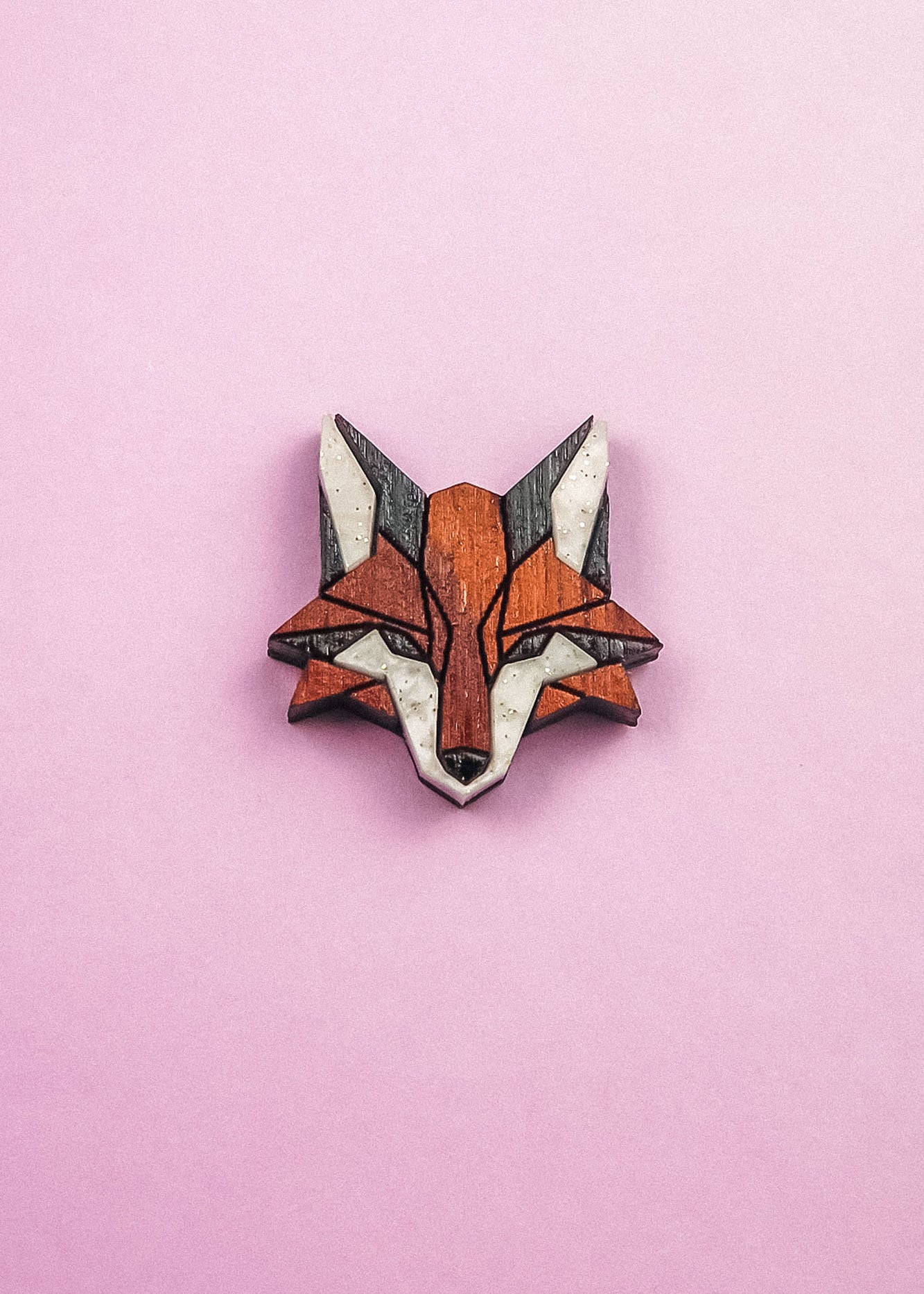 A geometric, stylized fox head brooch—handcrafted from recycled wood in shades of brown, black, and white—rests on a light pink background, making it an eco-conscious accessory with modern flair.