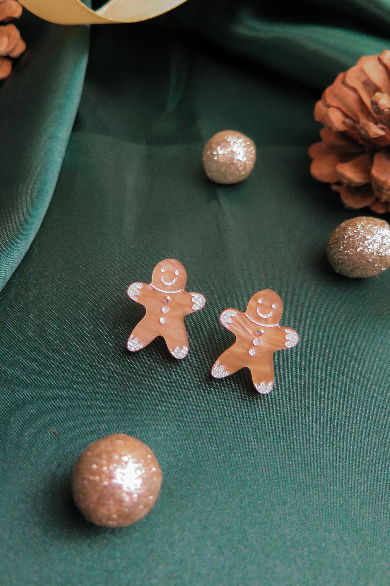 Two gingerbread man earrings rest on green fabric, surrounded by sparkling gold ornaments and a pinecone, making these festive accessories the perfect Christmas jewelry for any holiday look.