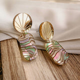 A pair of handcrafted gold earrings with fan-shaped tops and oval, iridescent abalone shell pendants rests on a light wood surface, with a white fabric backdrop in soft natural light.