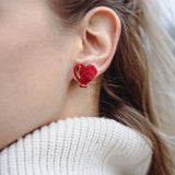 Close-up of a woman's ear wearing playful Heart Balloon Studs—red heart-shaped earrings with a small gold chain. Part of her face and blonde hair is visible, complemented by a white ribbed turtleneck sweater.