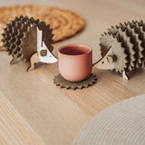 Two wooden hedgehog-shaped felt coasters hold a round, pink cup on a wooden table with woven placemats in the background—a charming touch of home decor.