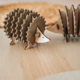 A small, decorative wooden hedgehog coaster holder made from layered, textured materials stands on a light wooden surface, with another similar figurine blurred in the background—perfect for adding charm to your home decor.