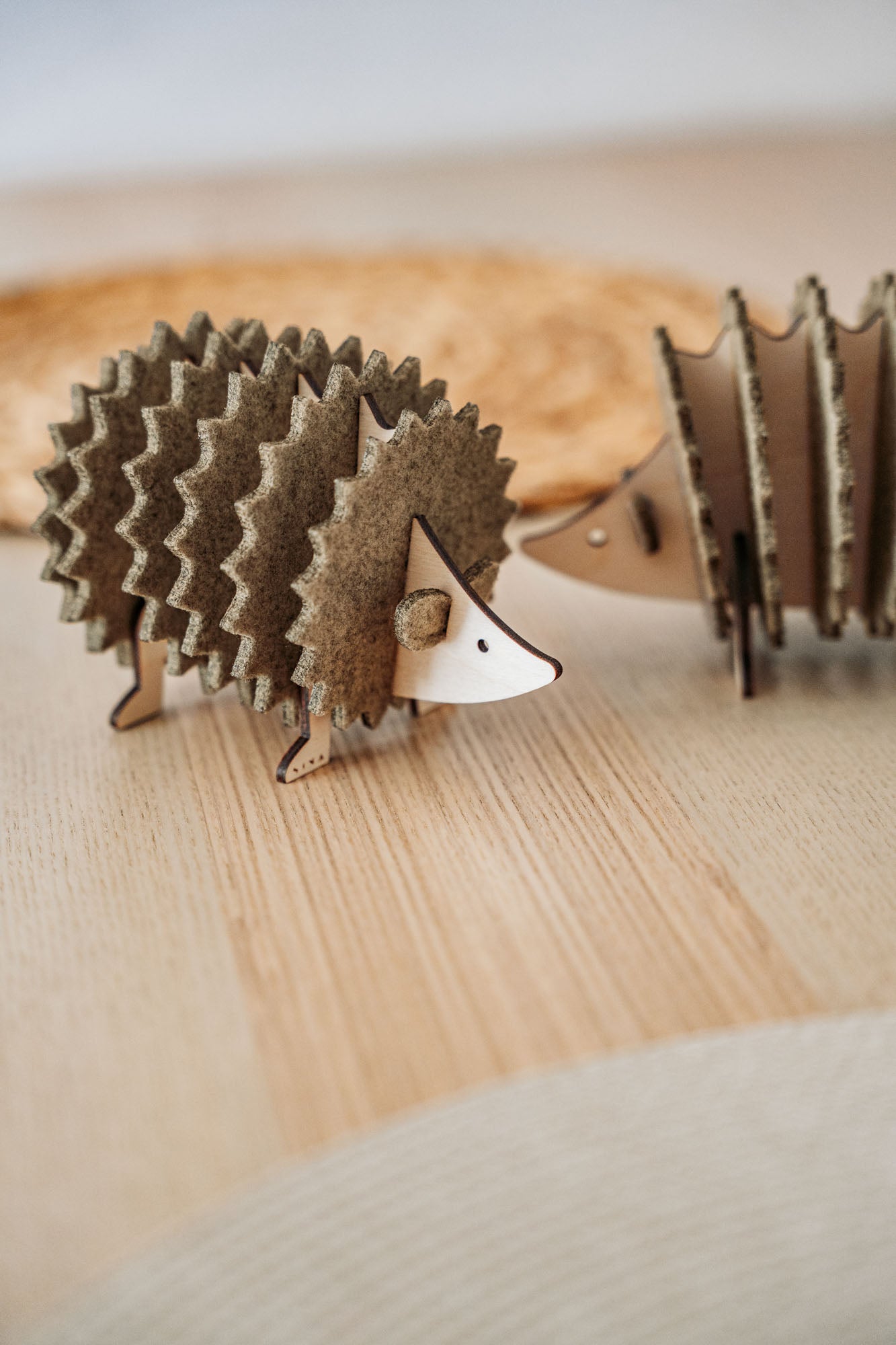 A small, decorative wooden hedgehog coaster holder made from layered, textured materials stands on a light wooden surface, with another similar figurine blurred in the background—perfect for adding charm to your home decor.