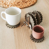 A white mug and a pink cup sit on spiky felt coasters shaped like a hedgehog, adding playful home decor flair to a light wooden table with a woven placemat in the background.