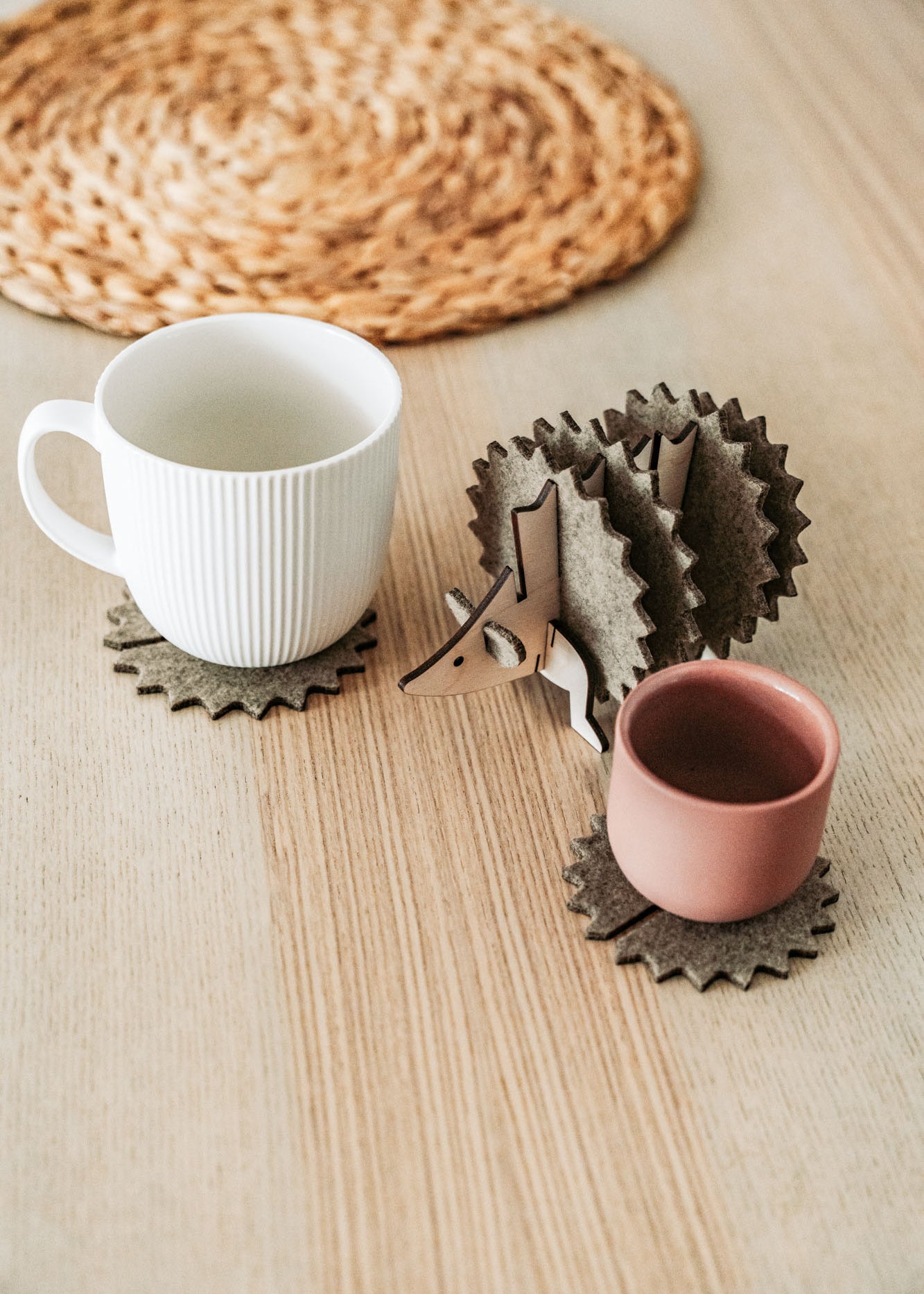A white mug and a pink cup sit on spiky felt coasters shaped like a hedgehog, adding playful home decor flair to a light wooden table with a woven placemat in the background.