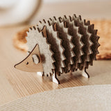 A charming wooden hedgehog coasters holder, perfect for home decor, sits on a light table with a woven placemat and blurred accents in the background.