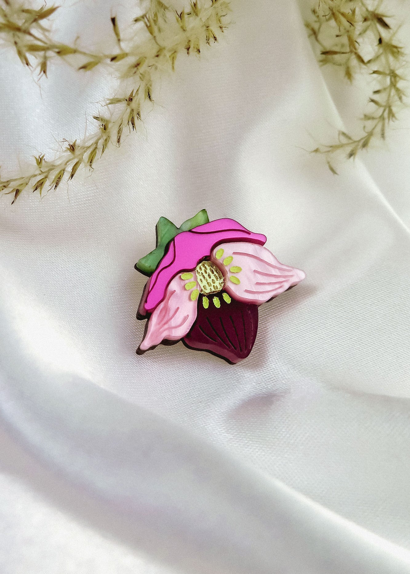 A handmade Purple Hellebore Flower lapel pin rests on white satin fabric, with delicate sprigs of dried grass in the background.