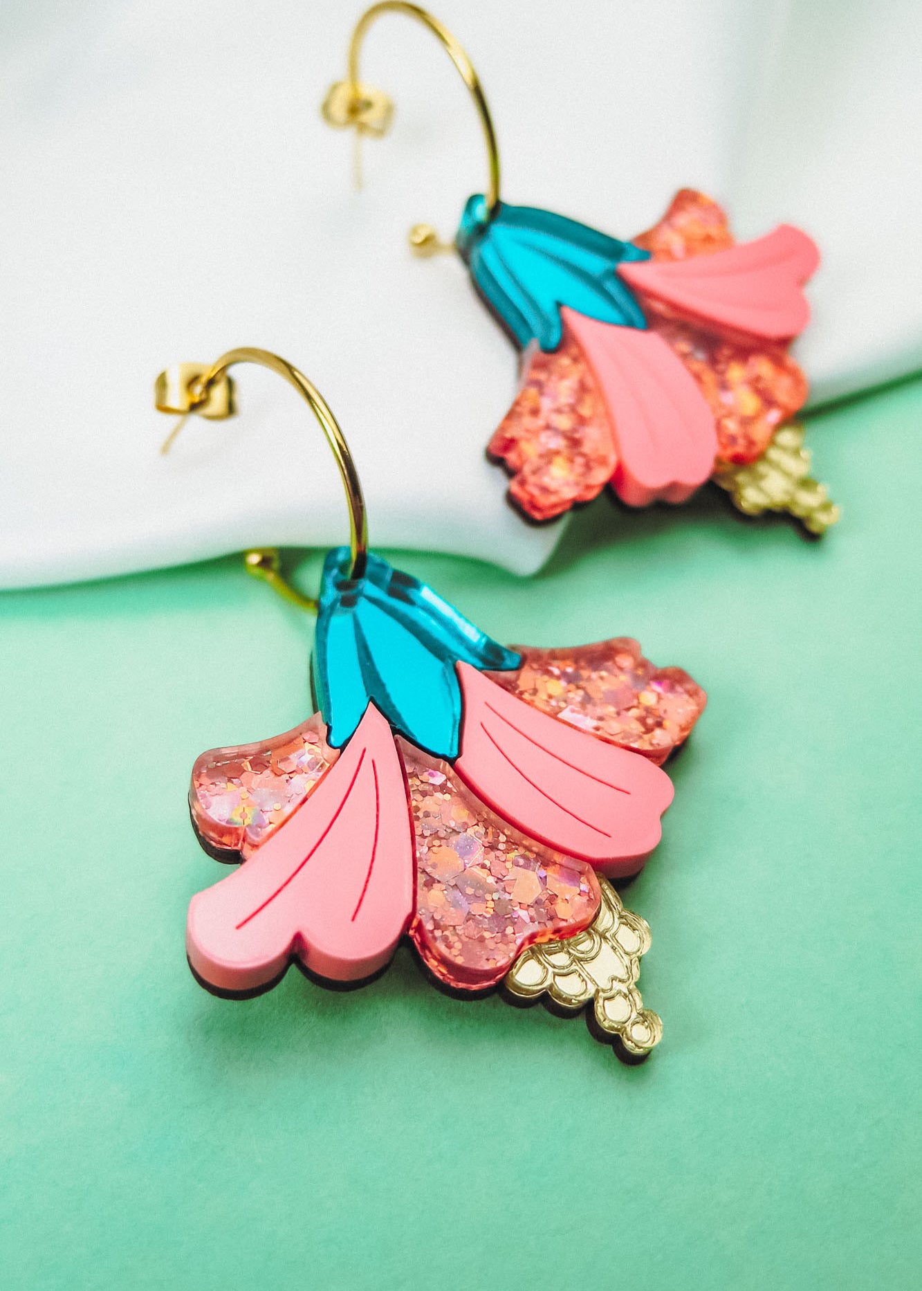 Vibrant Hibiscus Earrings with gold hooks, featuring colorful pink, blue, and glittery accents, make a bold tropical statement against a striking green and white background.