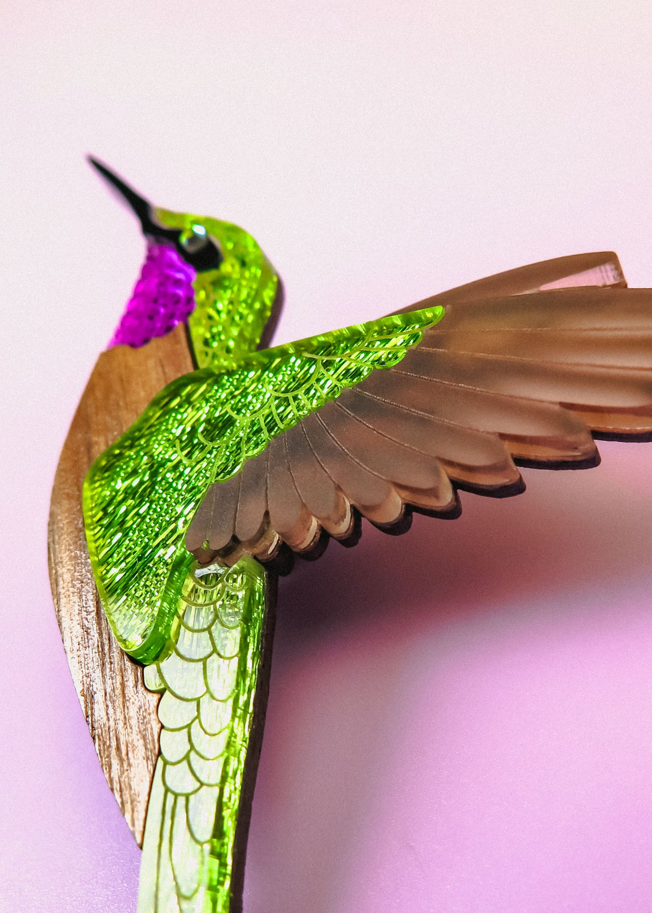 A close-up of a handcrafted hummingbird brooch in vibrant colors—metallic green and purple with brown wings—set against a soft pink background.