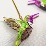 Close-up of handcrafted Hummingbird Earrings featuring gold, green, purple, and white geometric patterns, displayed on a white fabric background—a vibrant celebration of sustainable craftsmanship.