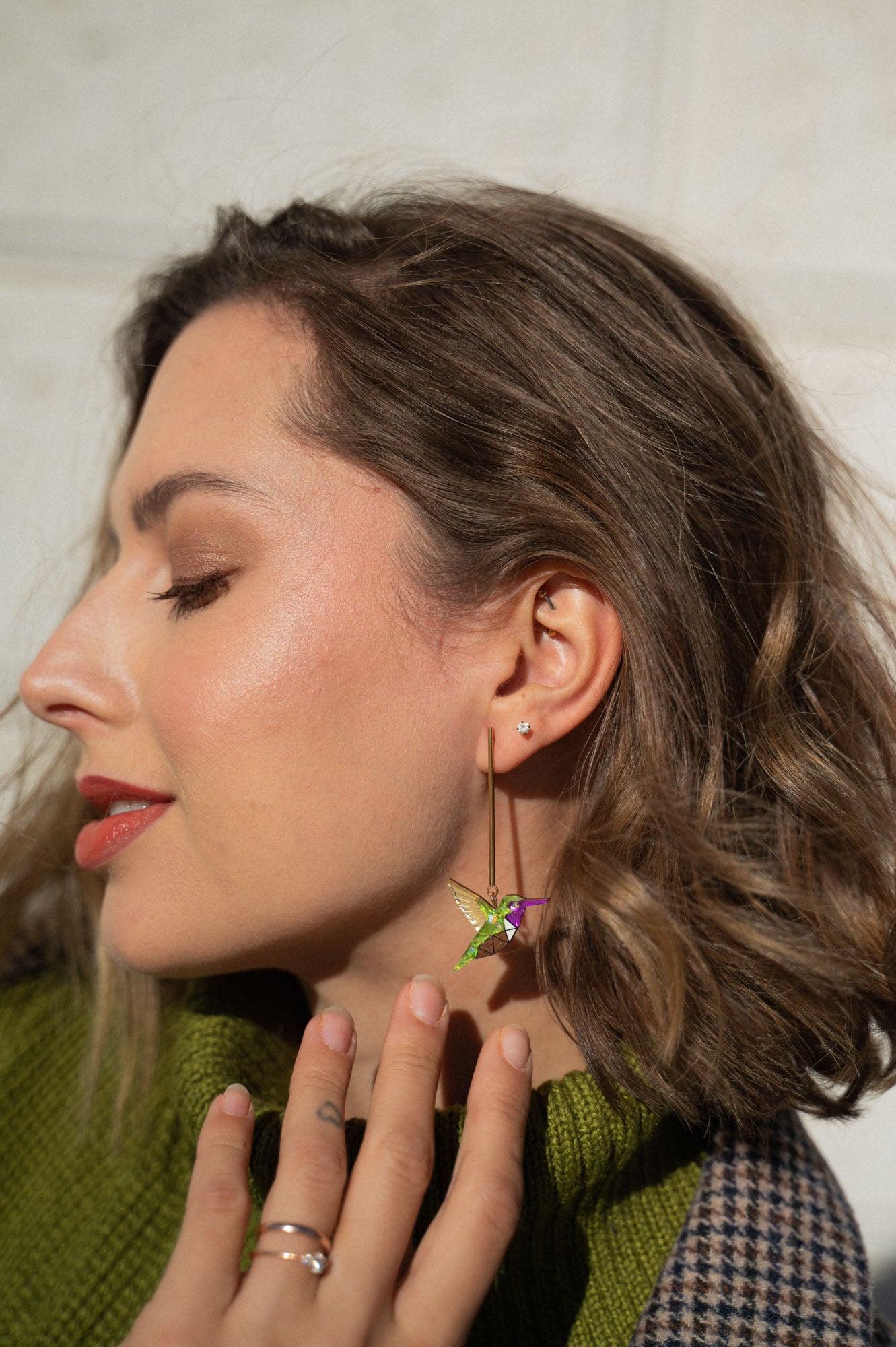 A woman with wavy brown hair and peach-toned makeup wears a green sweater and handcrafted Hummingbird Earrings. She smiles softly with her eyes closed, touching her neck as she showcases sustainable craftsmanship.