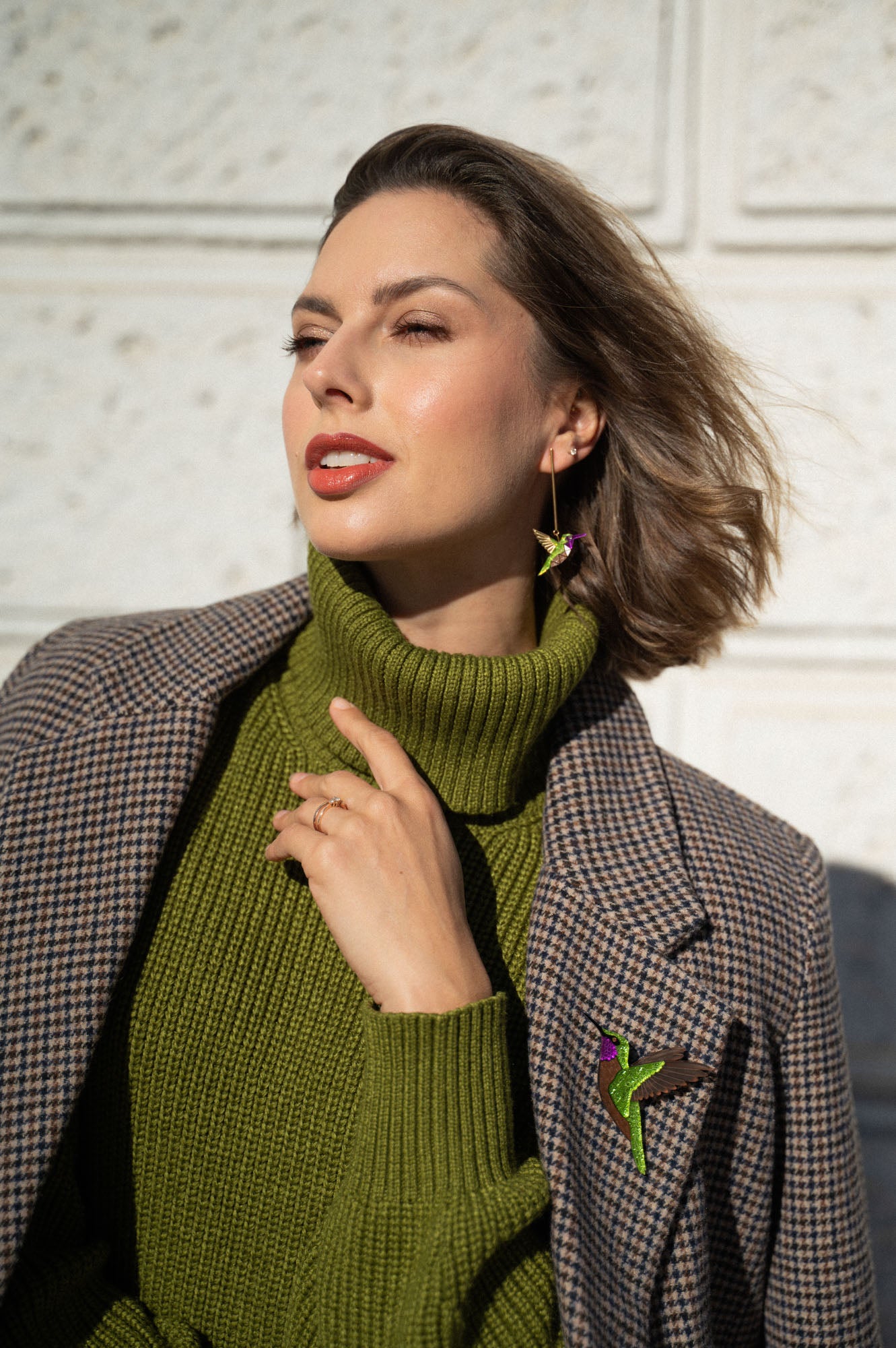 Woman wearing a green turtleneck sweater and plaid coat, accessorized with handcrafted Hummingbird Earrings and a brooch, poses outdoors against a light textured wall, touching her collar and looking off to the side.