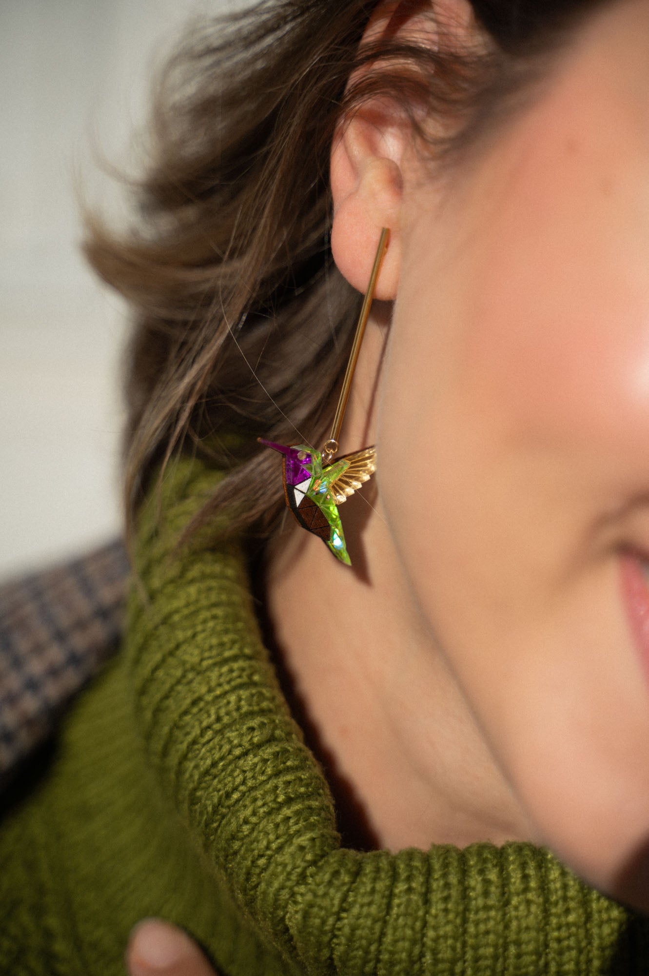 Close-up of a woman's ear wearing a long gold Hummingbird Earring, handcrafted with green, gold, and purple details. She pairs it with a green knitted turtleneck sweater, showcasing sustainable craftsmanship.
