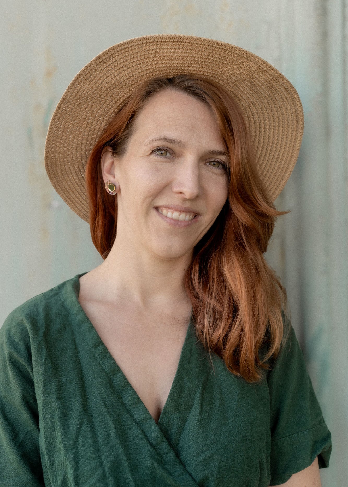 A woman with long auburn hair wears minimalist geometric stud earrings, a wide-brimmed straw hat, and a green dress, smiling softly while standing in front of a neutral-colored wall.